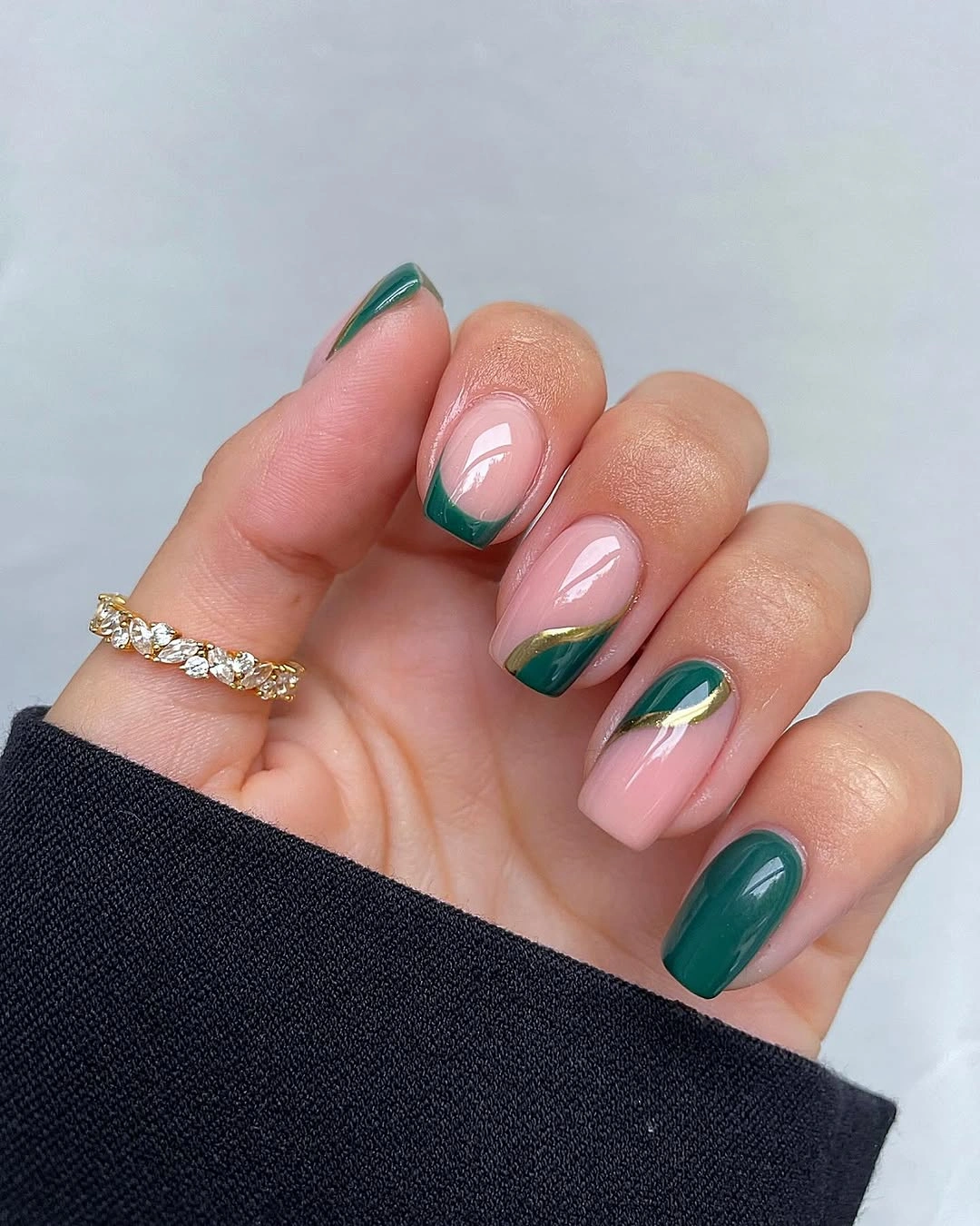 Pale pink almond nails with deep green French tips and curved gold accents.
